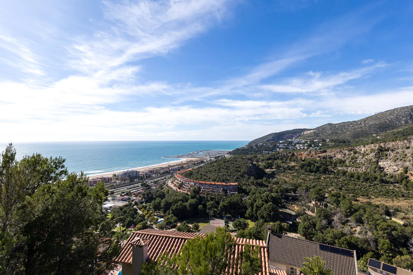 Scenic view of a coastline with a sandy beach, blue ocean, and green hills under a partly cloudy sky. Red-tiled roofs of houses are visible in the foreground.