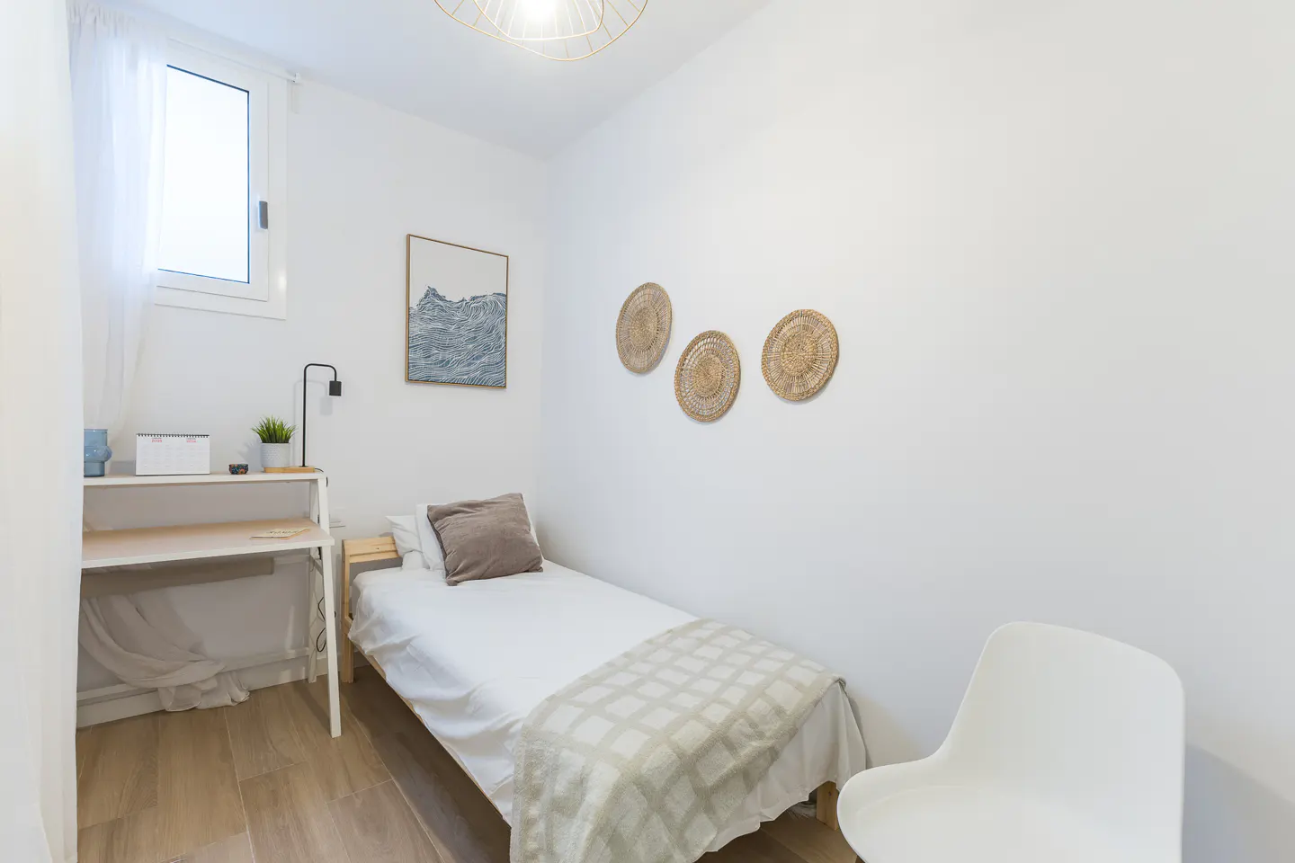 Bright, minimalist bedroom with a single bed, desk, and white chair. Woven wall decor and a framed print add subtle texture.