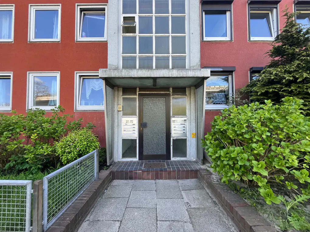 Apartment building entrance with a gray door, mailboxes, and a concrete awning. The building is red with white-framed windows and green bushes.