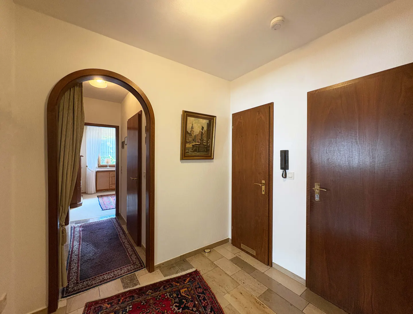 Hallway with arched doorway, wood doors, and stone tile floor. A painting hangs on the white wall. Rugs add color to the floor.
