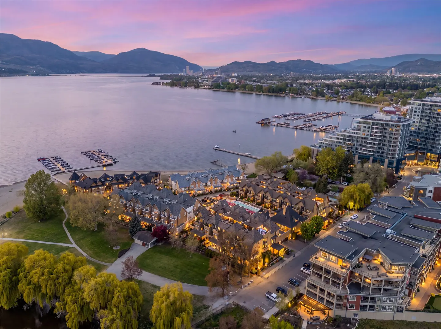 Aerial view of waterfront condos and townhouses at dusk, with mountains, lake, and marina in the background.