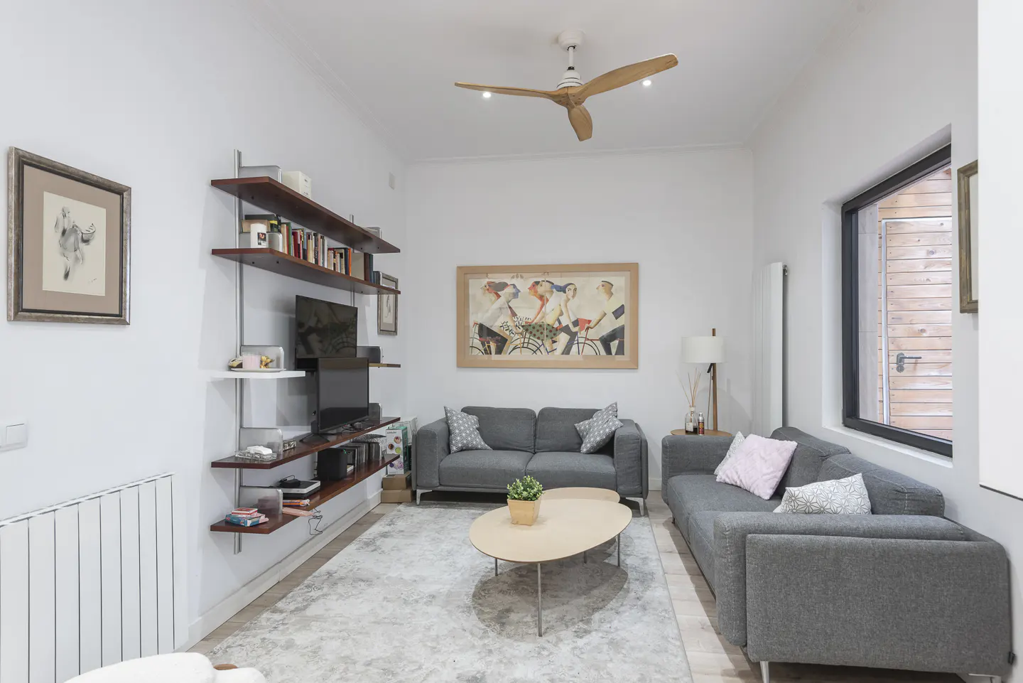 A bright living room with gray sofas, a light wood coffee table, and a wooden ceiling fan. Shelves with books and a TV are on the left wall.
