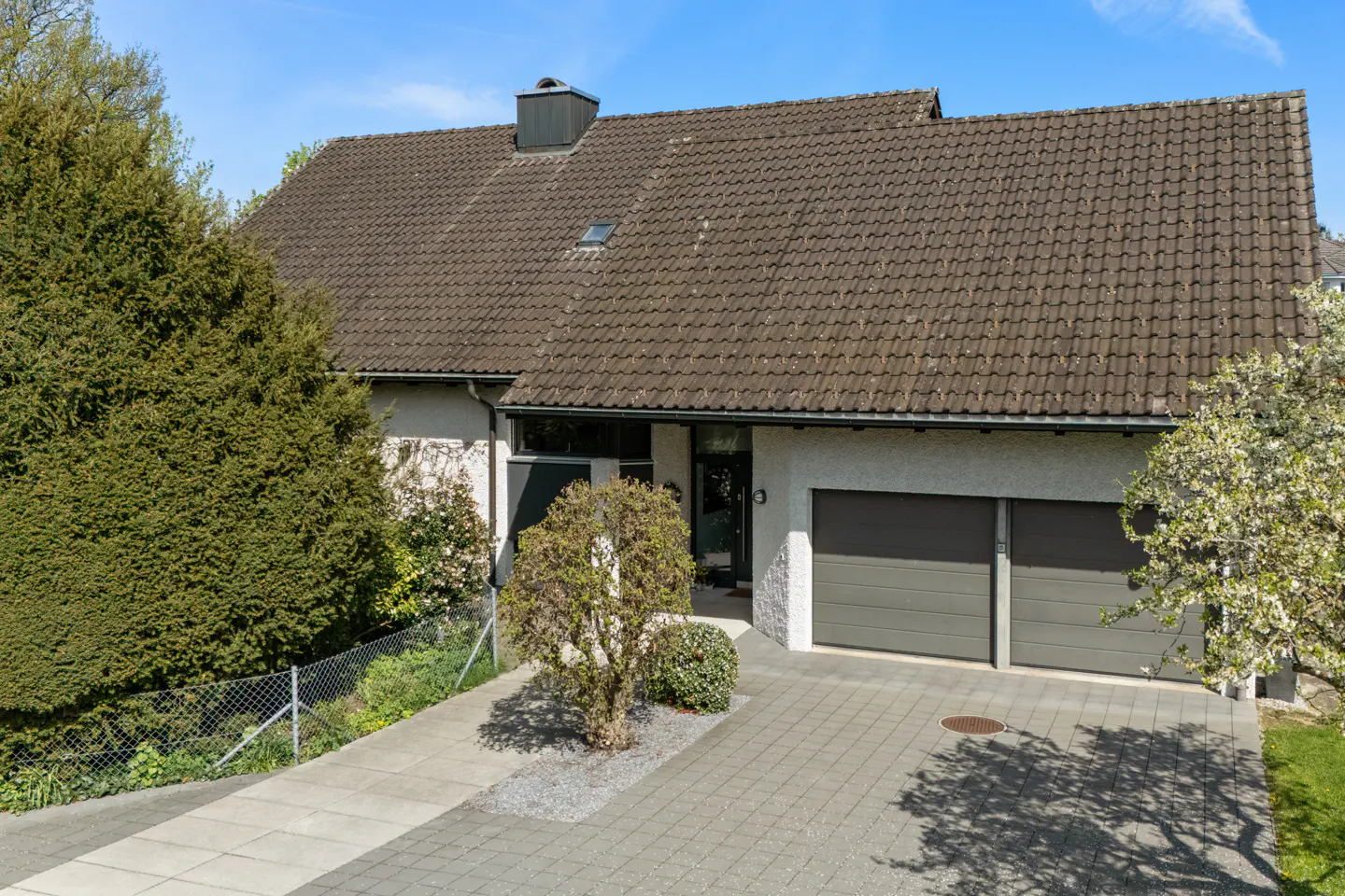 Exterior of a one-story house with a brown tiled roof, gray garage doors, and a paved driveway. Green trees and bushes surround the house.