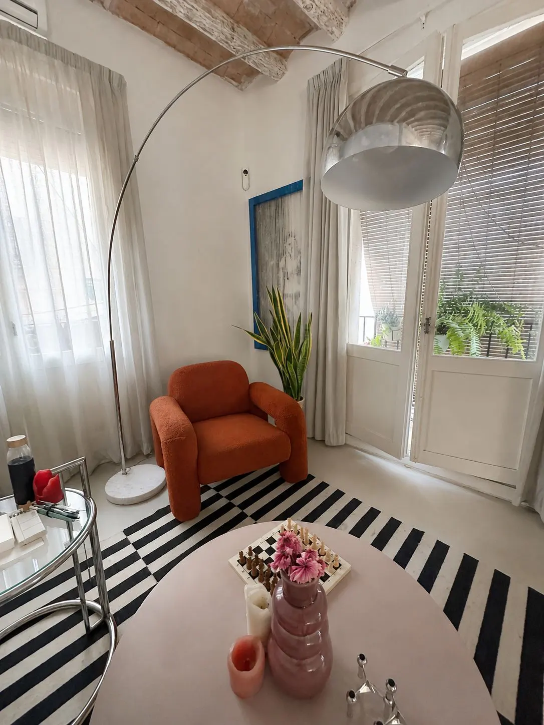 A cozy living room with a burnt orange armchair, a large arched floor lamp, and a round table with a chess set and flowers.