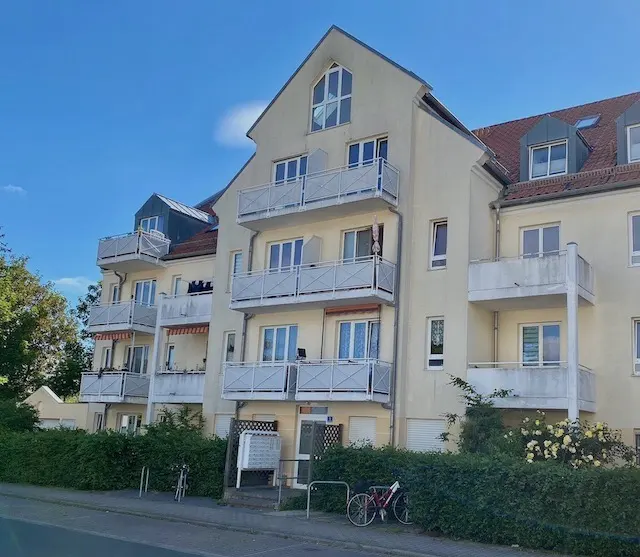 A three-story apartment building with white balconies and a light yellow facade under a blue sky.