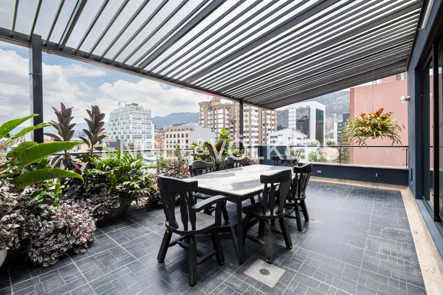 Outdoor patio with black table and chairs, plants, and a city view under a slatted pergola.
