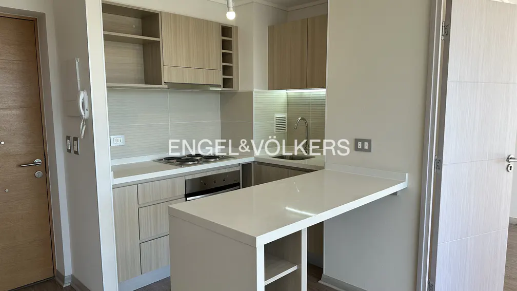 A modern kitchen with light wood cabinets, white countertops, and stainless steel appliances. A breakfast bar extends from the counter.