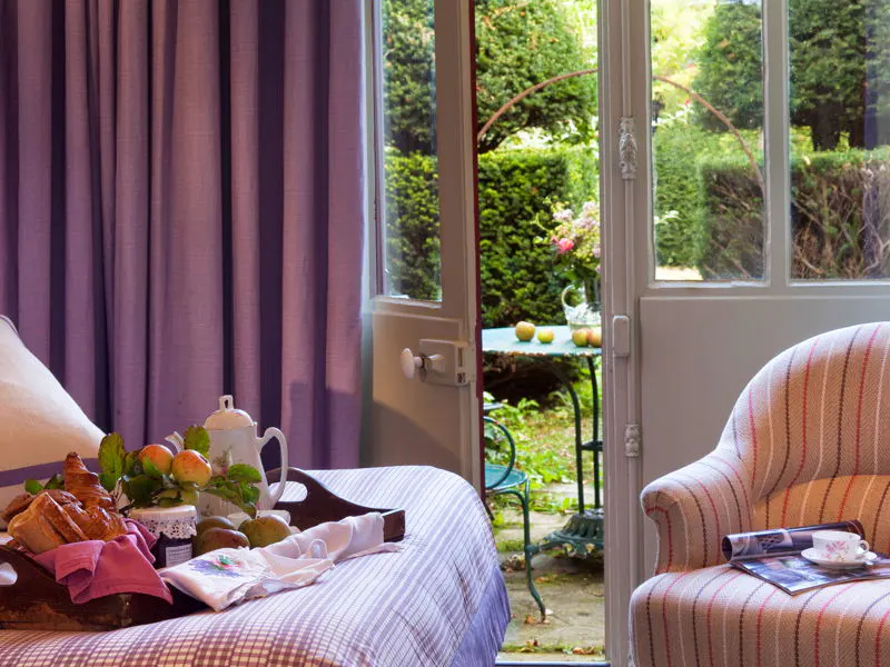 Cozy bedroom with breakfast tray on bed, open door to garden. Striped armchair with magazine and teacup. Lavender curtains.