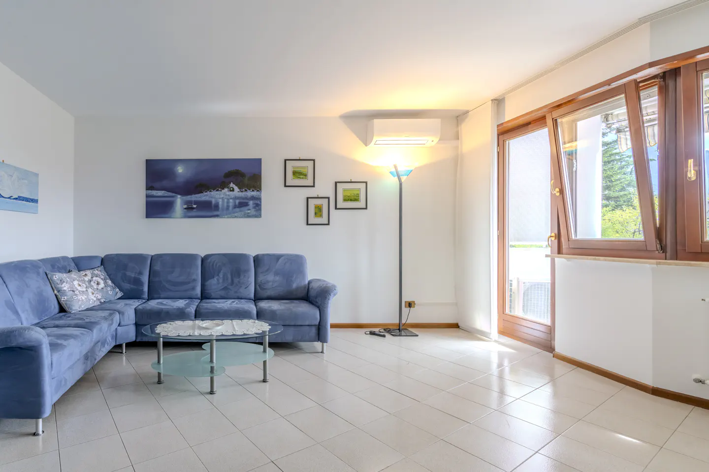 Bright living room with a blue sectional sofa, glass coffee table, and white tile floor. Artwork hangs on the white walls, and a tall window lets in natural light.