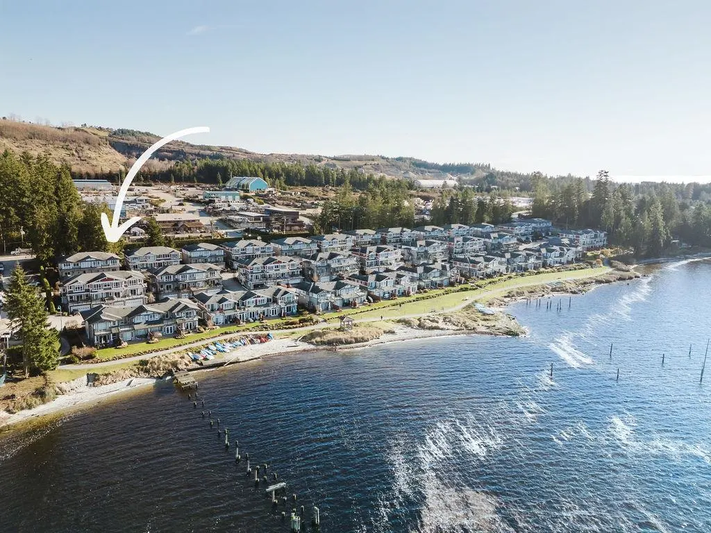 Aerial view of coastal townhouses with an arrow pointing to a specific unit near the water's edge. Blue water and green trees surround the property.