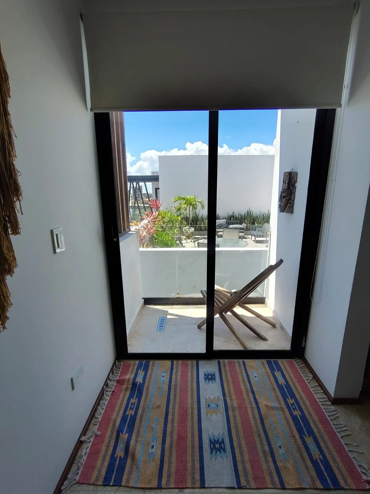 View from a room with a colorful rug, through a sliding glass door to a balcony with a wooden lounge chair and a garden.