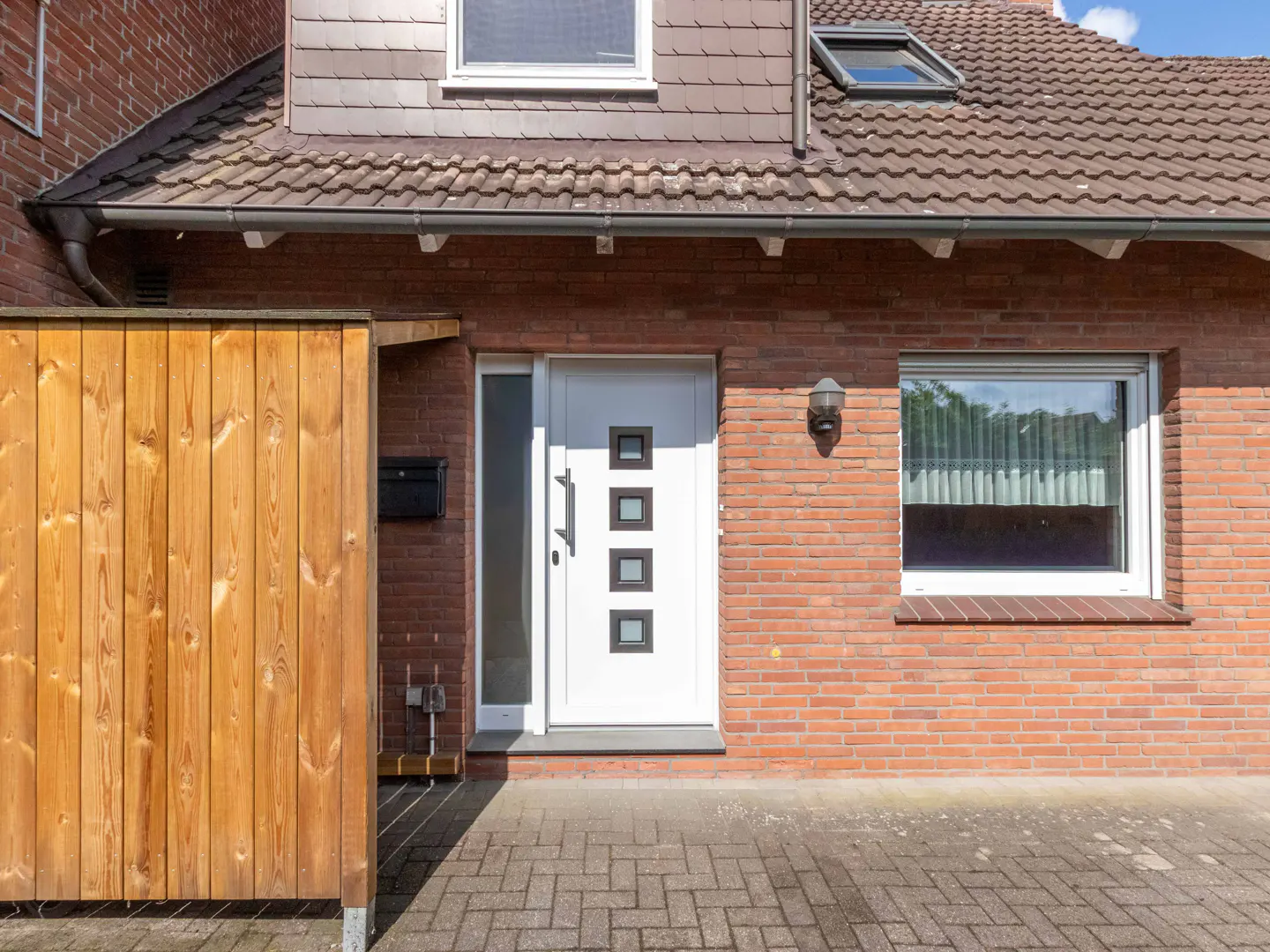 Exterior of a brick house with a white front door with square windows, a wooden fence, and a brick walkway.