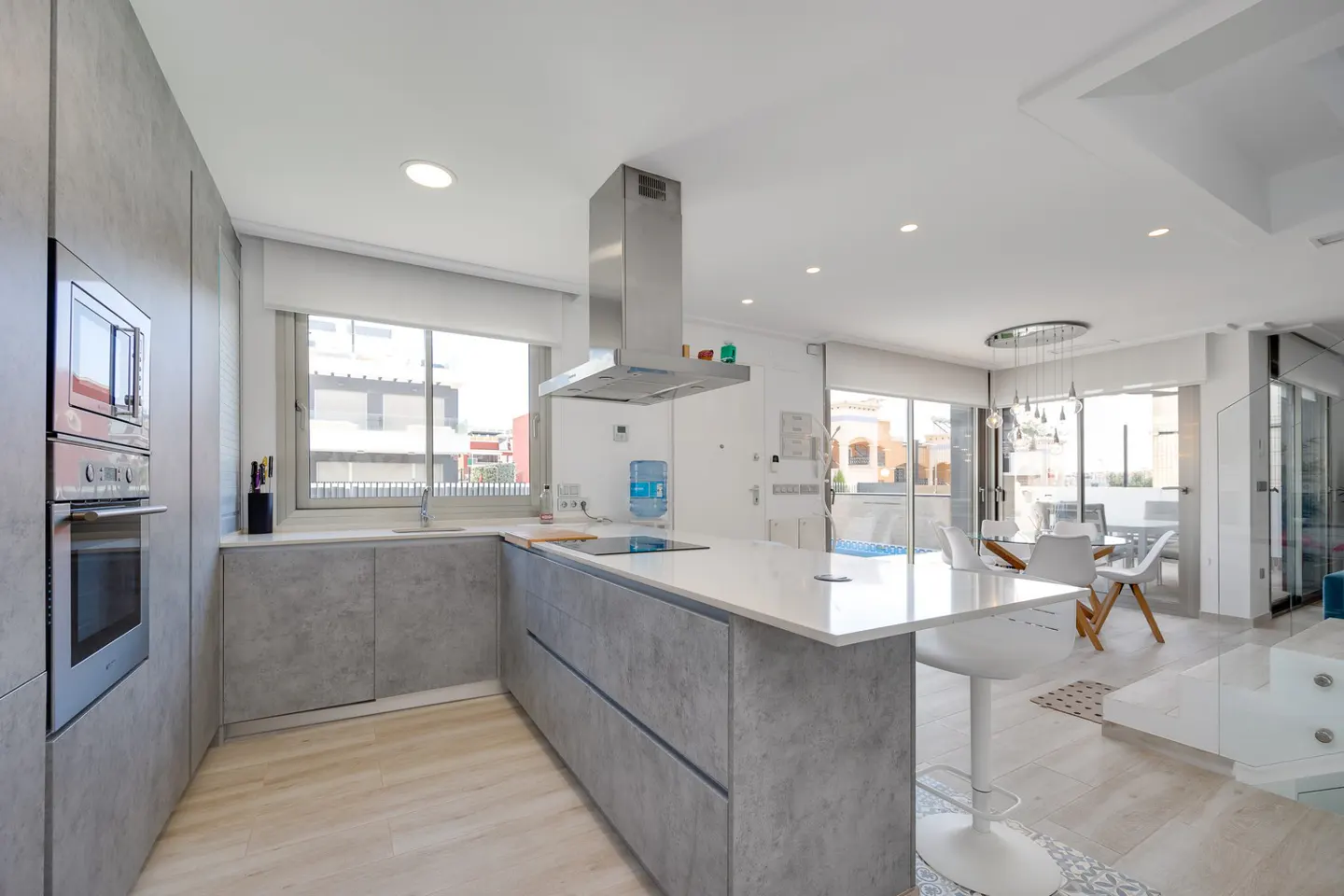 Modern kitchen with grey cabinets, white countertops, stainless steel hood, and a breakfast bar with white stools.