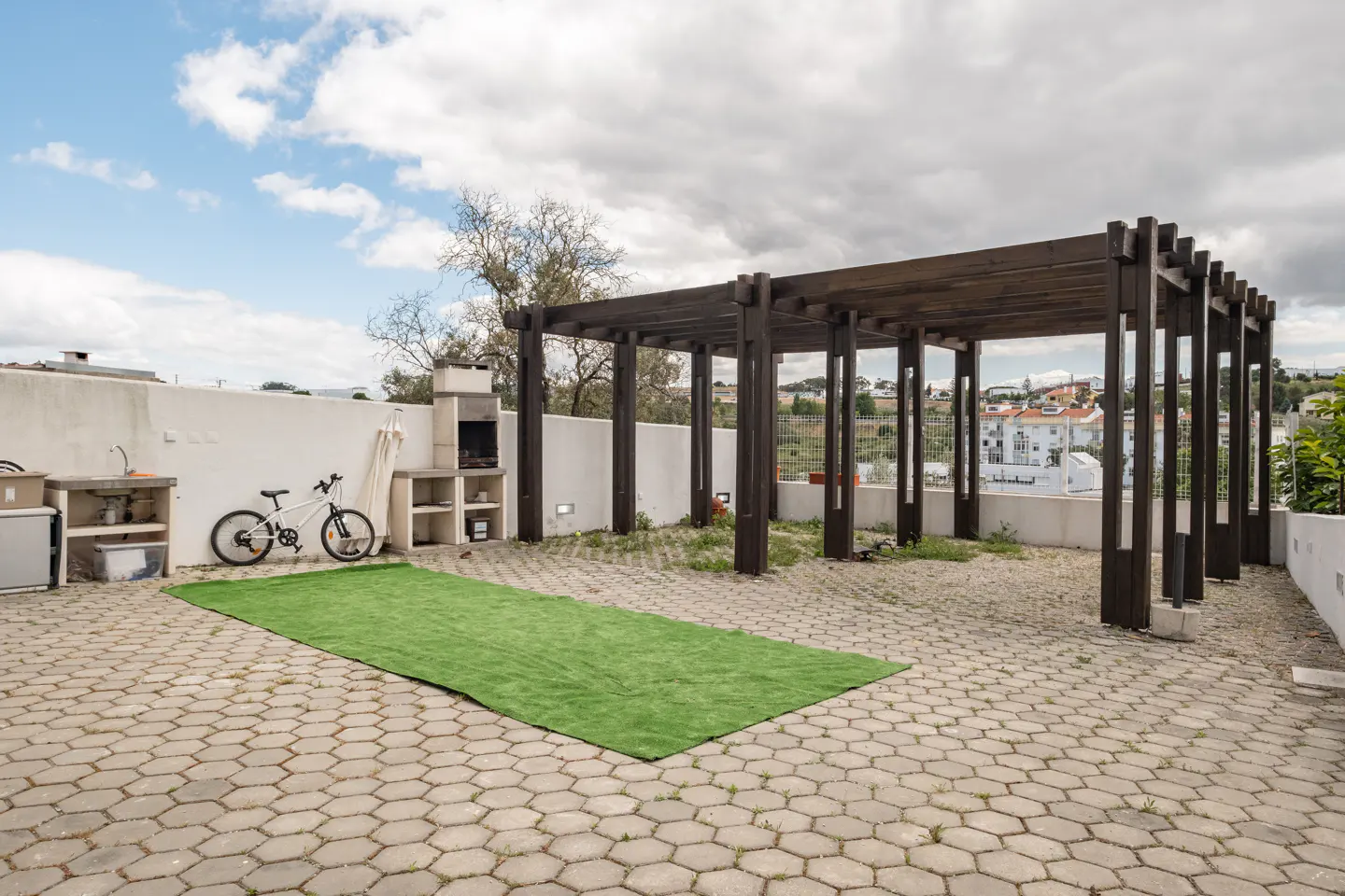 Outdoor patio with a dark wood pergola, a built-in grill, a sink, a bicycle, and a green rug on a gray stone floor.