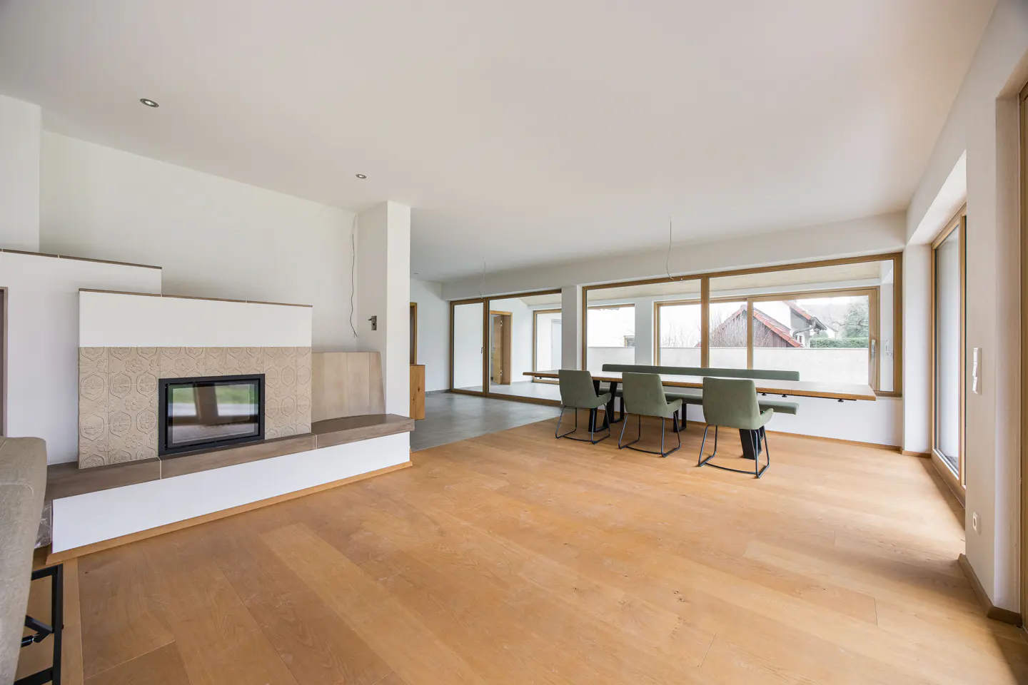 Bright, open-plan living space with light wood floors, a fireplace, and a dining table with green chairs and a bench by large windows.