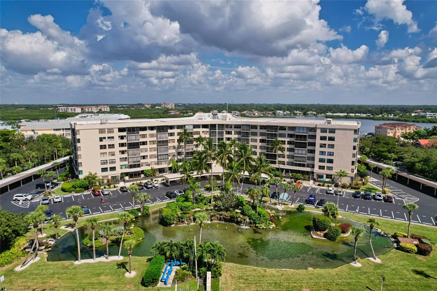 Aerial view of a beige apartment complex with a central pond, palm trees, and parking lot under a blue sky with clouds.