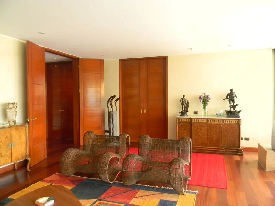 Living room with two wicker chairs on a colorful rug, wooden floors, and cabinets with sculptures.