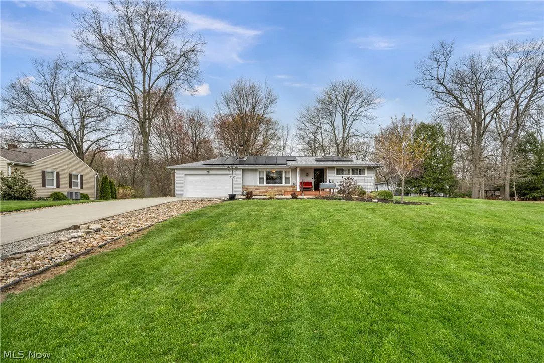 A single-story gray house with a green lawn and a driveway leading to a white garage. Trees are in the background.