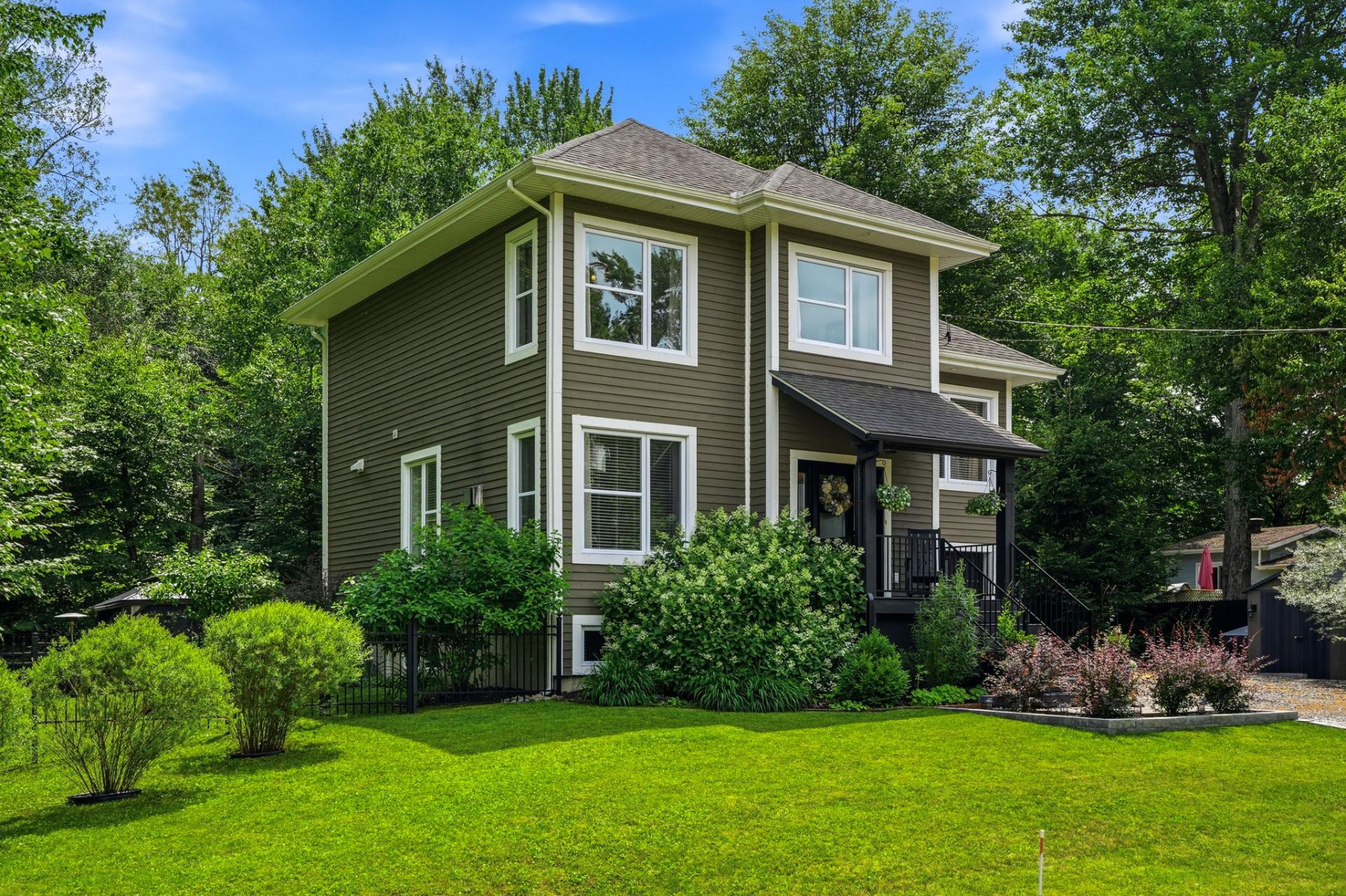 Two-story olive green house with white trim, a covered front porch, and a lush green lawn surrounded by mature trees. [AI-generated text]