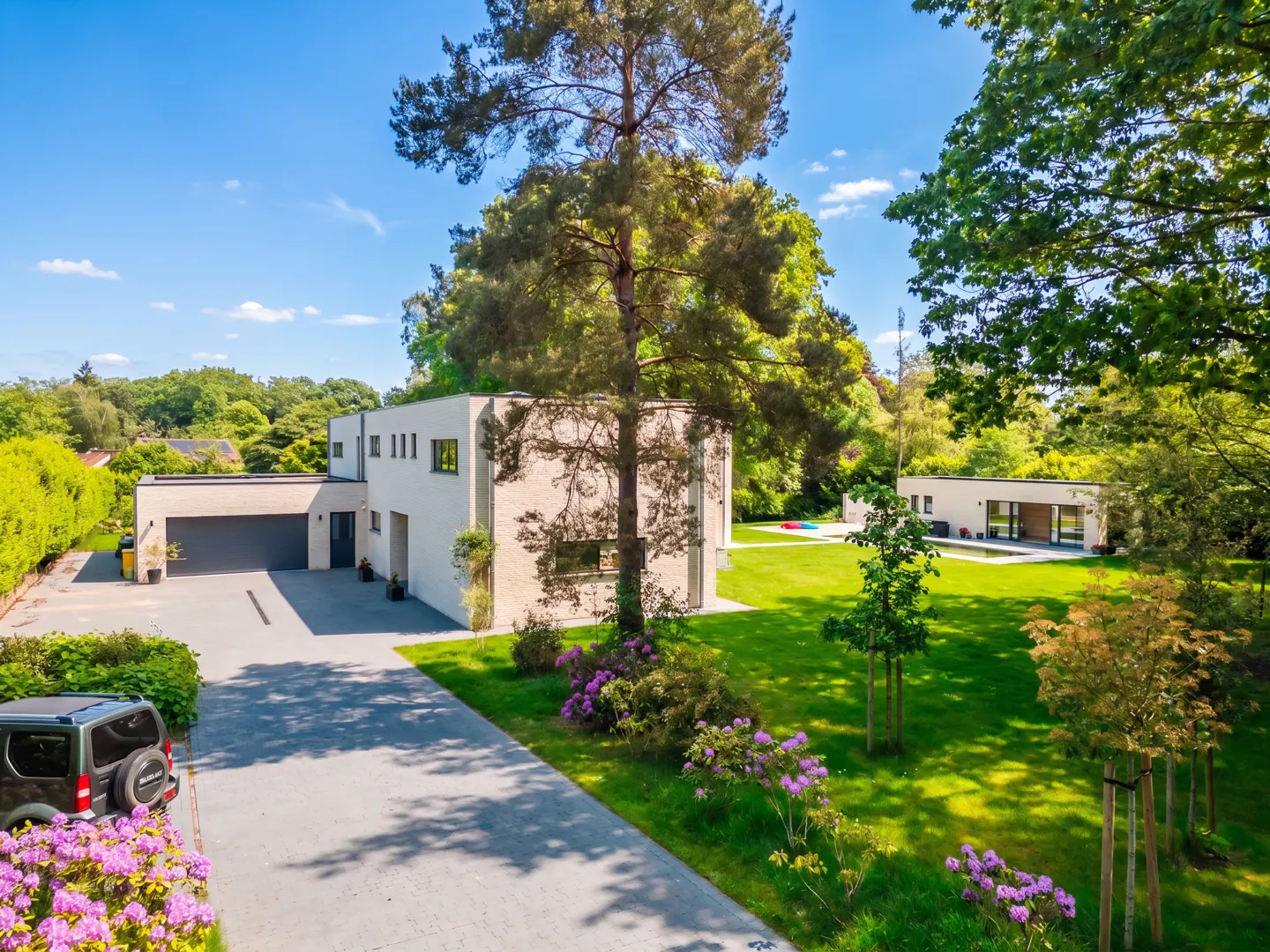 Modern white house with a gray driveway, green lawn, trees, and purple flowers under a blue sky.