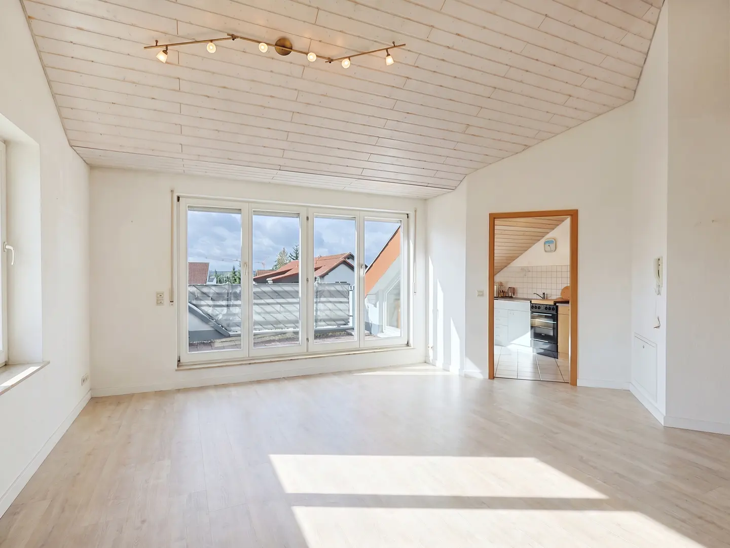Bright, empty room with white walls, wood floors, and a wood-paneled ceiling. Large windows overlook a balcony and neighborhood. A doorway leads to a kitchen.
