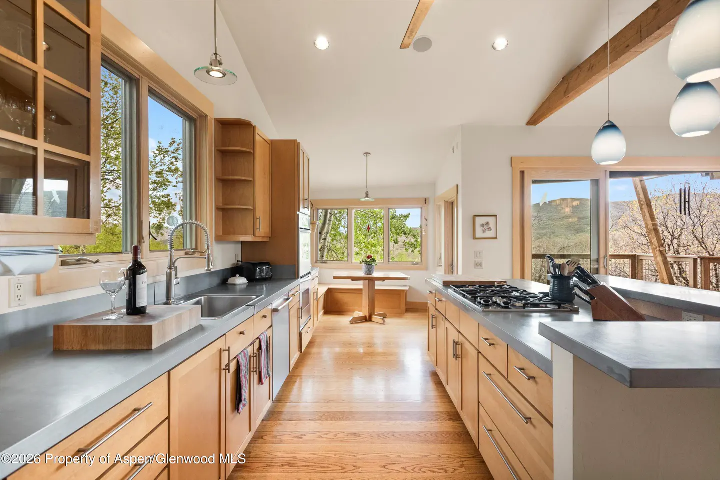 A bright kitchen with wood cabinets, gray countertops, and hardwood floors. A wine bottle sits by the sink, and a breakfast nook is visible in the background.
