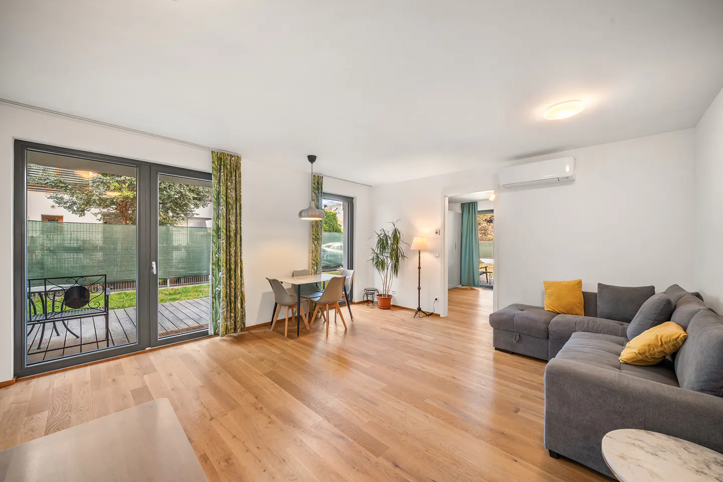 Bright living room with hardwood floors, gray sectional sofa with yellow pillows, dining table, and sliding glass doors to a patio.