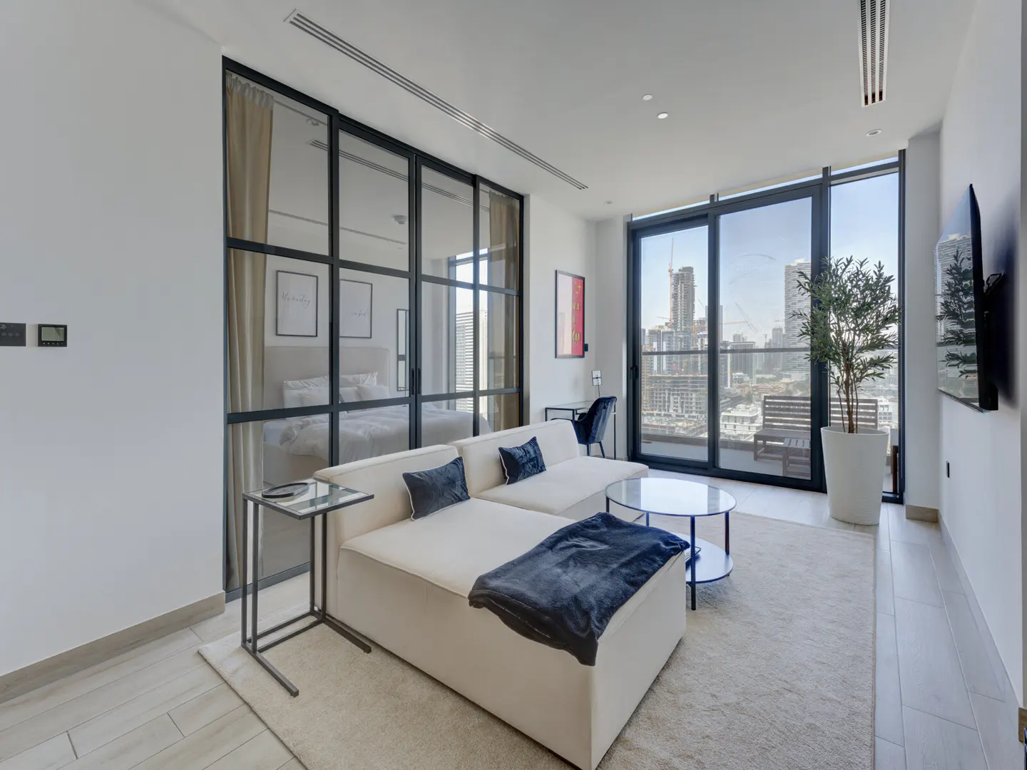 Living room with a white sofa, blue throw, and pillows. A glass wall separates the bedroom. City view through a large window.