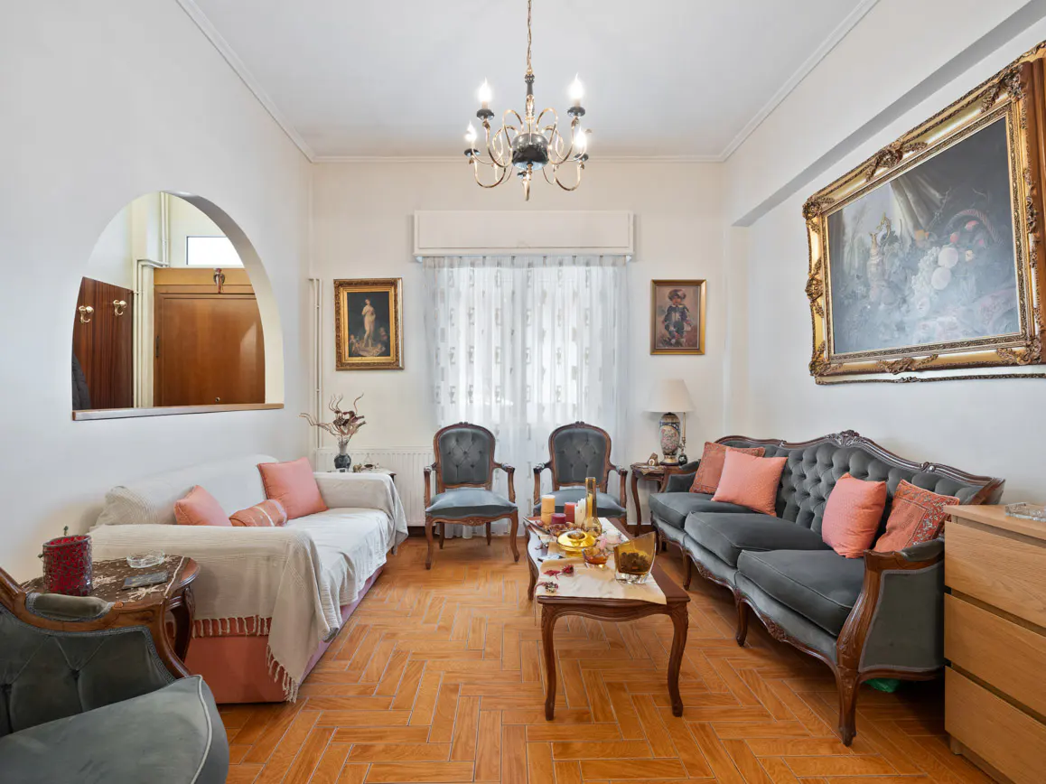 Living room with herringbone wood floors, white walls, and a mix of antique furniture, including a grey sofa and chairs.