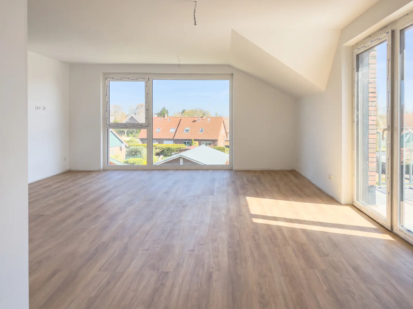 Bright, empty room with light wood floors, white walls, and large windows overlooking houses. Sunlight streams across the floor.