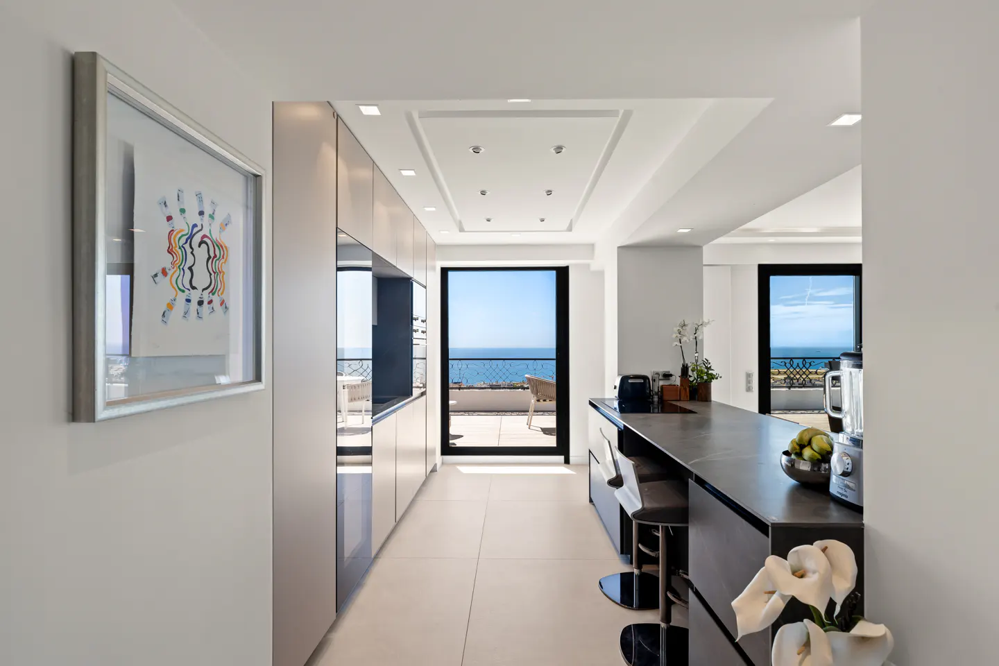Modern kitchen with dark island, bar stools, and floor-to-ceiling windows overlooking a sunny ocean balcony.