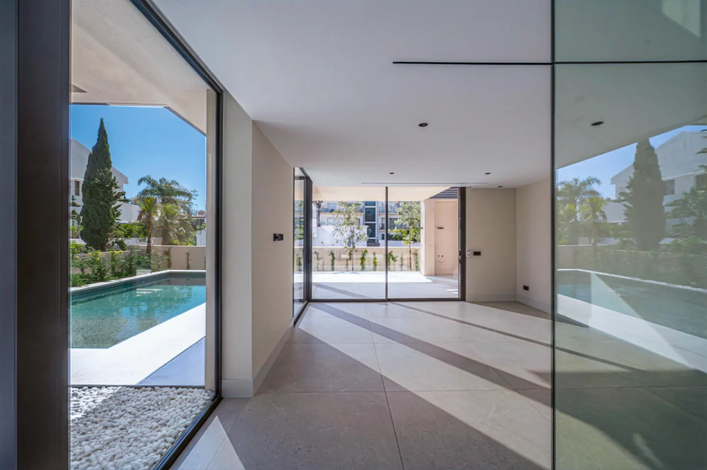 Modern, empty room with large windows. Outside, a pool and greenery are visible. The floor is light gray tile.
