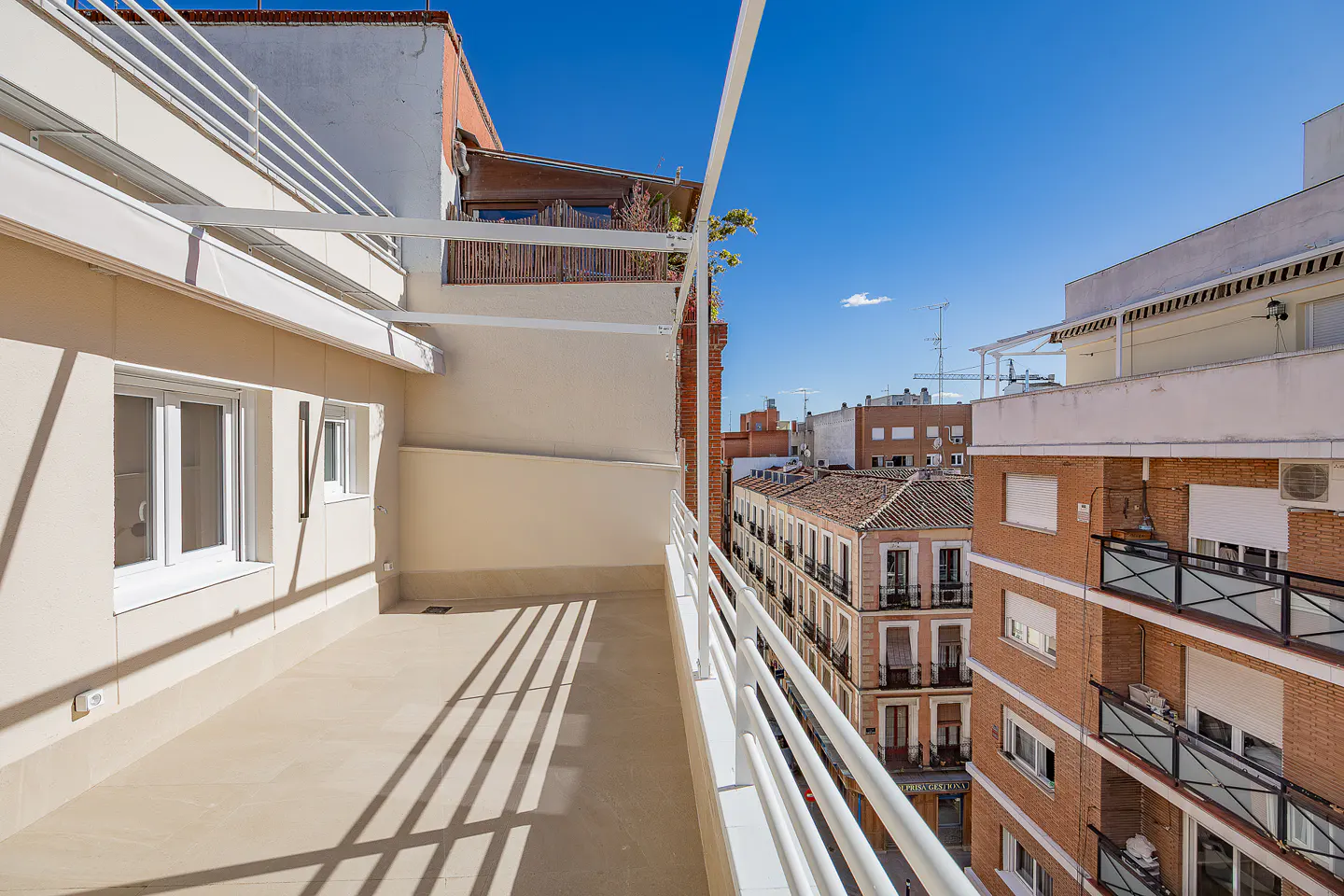 Balcony view of city buildings under a blue sky. White railings line the balcony edge. Cream-colored building with windows on the left.