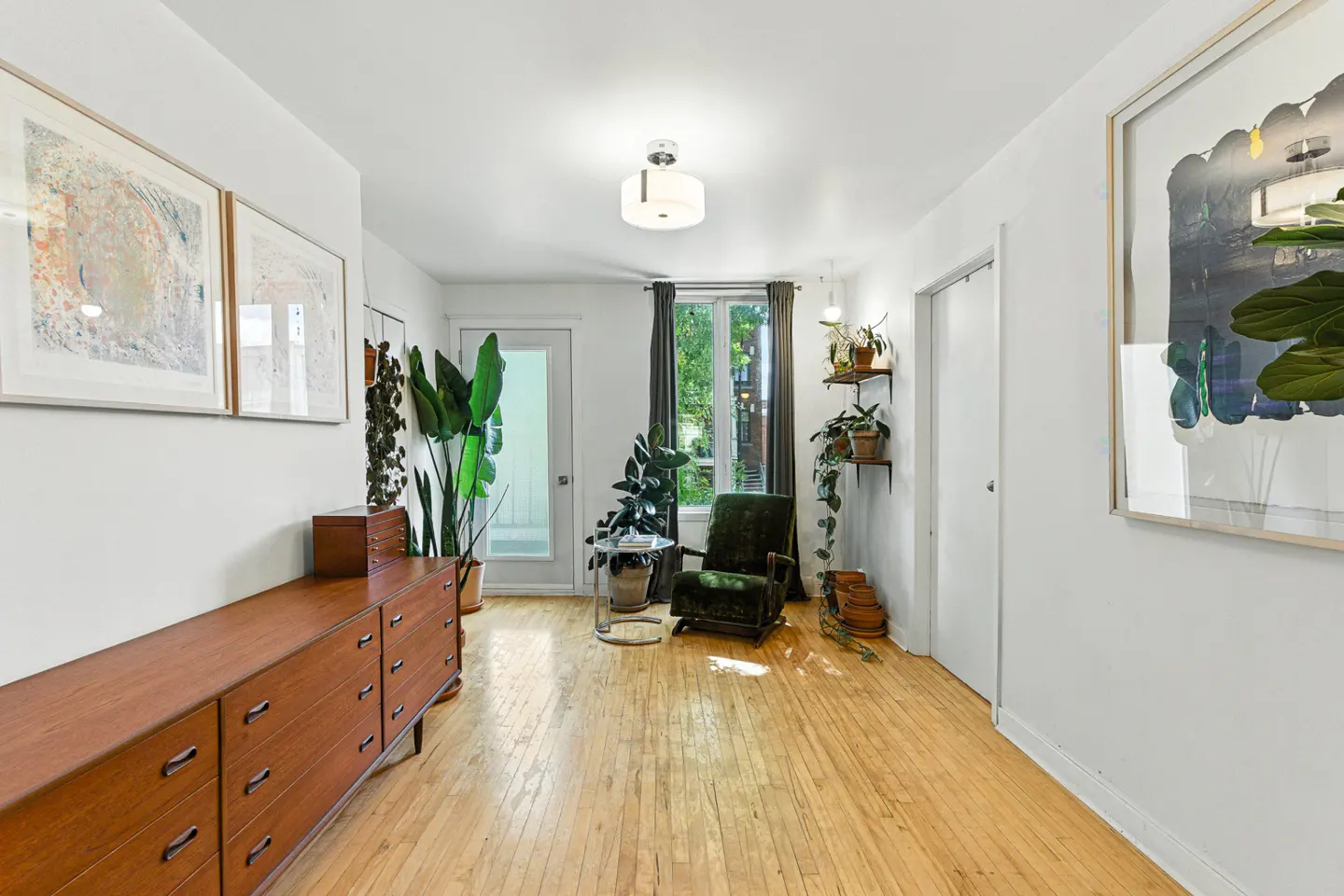 Bright living room with wood floors, white walls, and plants. A brown dresser sits under framed art. A green chair faces a window with gray curtains.