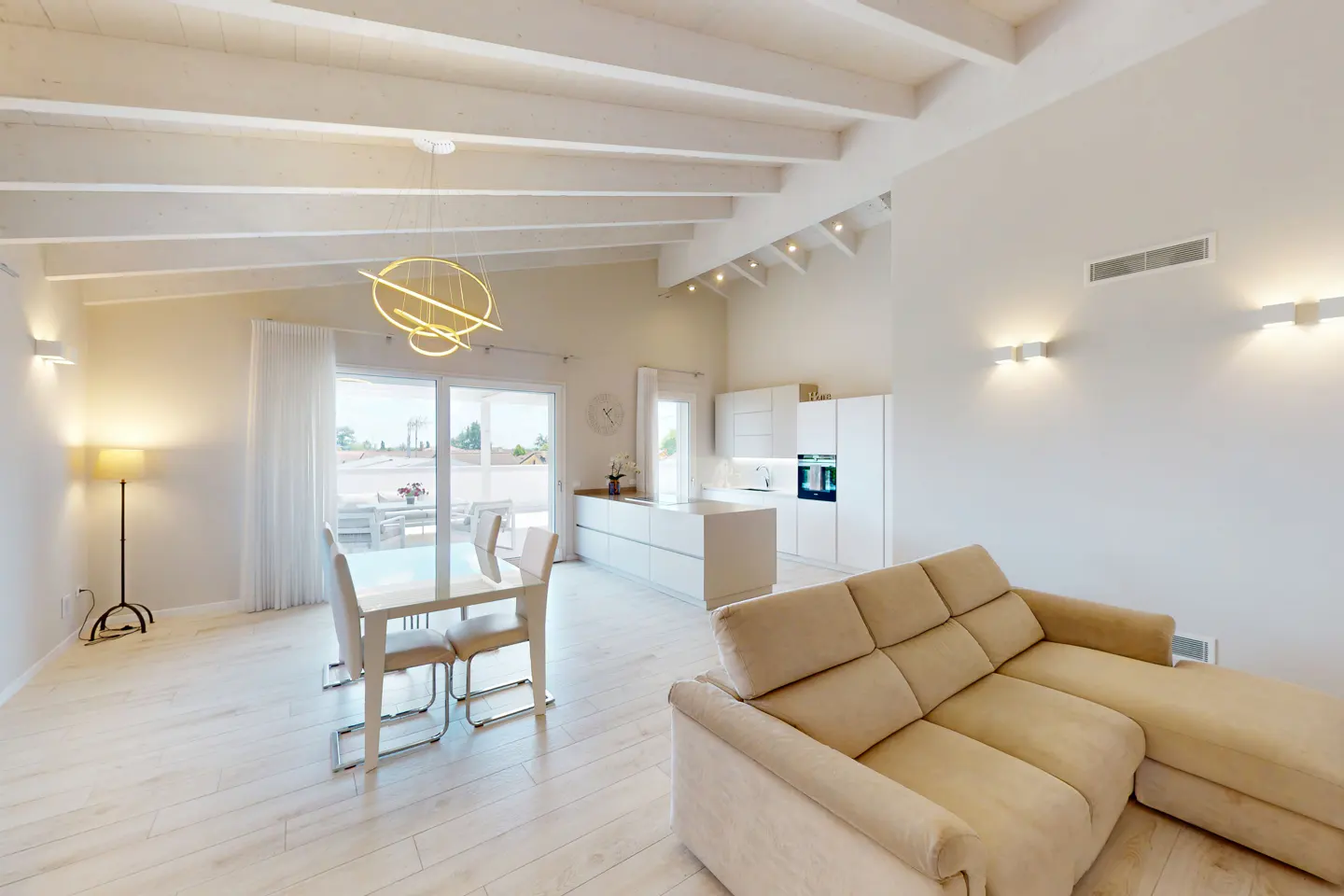 Bright, open-plan living space with white walls, wooden floors, and exposed beams. A beige sofa sits near a white kitchen and dining area with a modern chandelier.