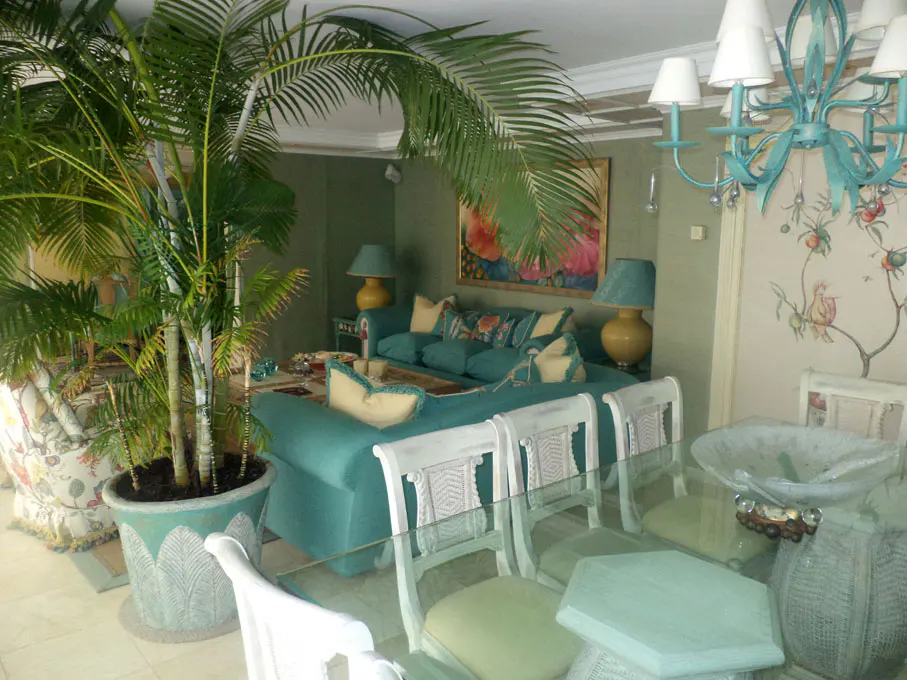 Living room with a turquoise sofa, white chairs, and a glass table. A large palm tree sits in a green pot. A turquoise chandelier hangs above.