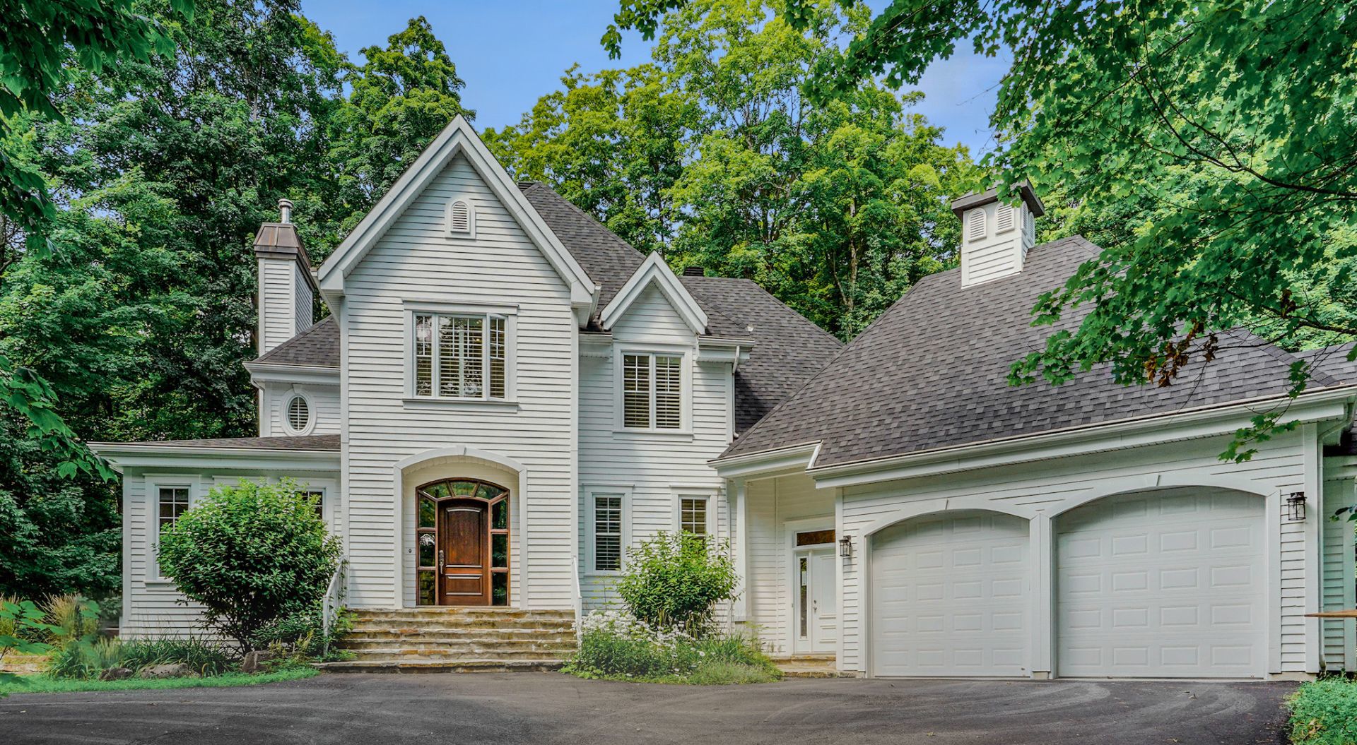 Exterior view of a two-story white house with a gray roof and a two-car garage, surrounded by green trees.