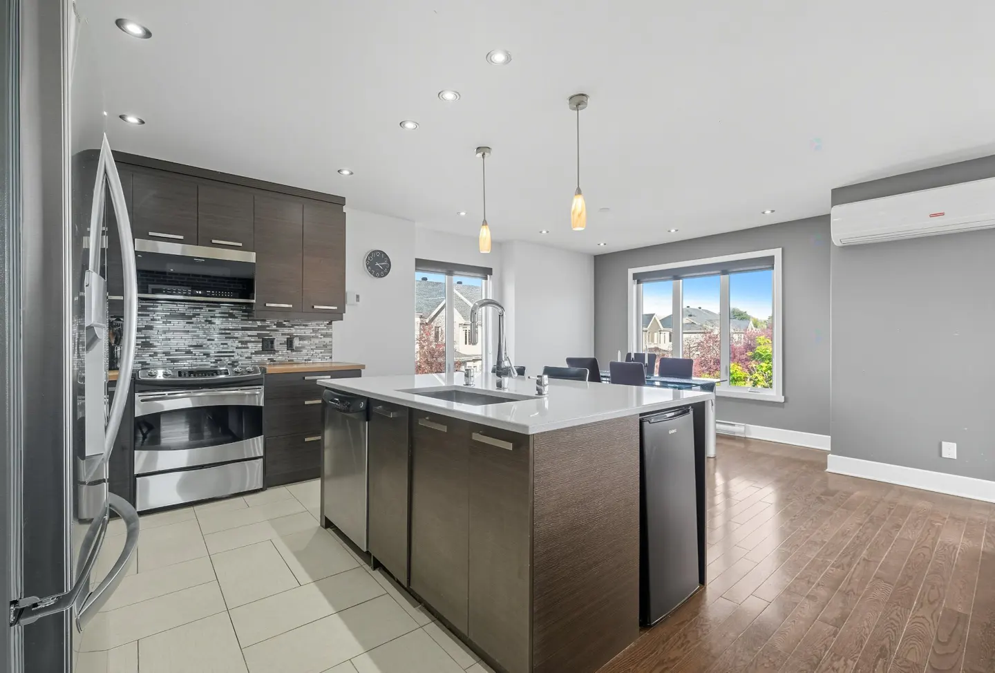 A modern kitchen with dark wood cabinets, stainless steel appliances, and a white countertop island. A dining area is visible in the background.
