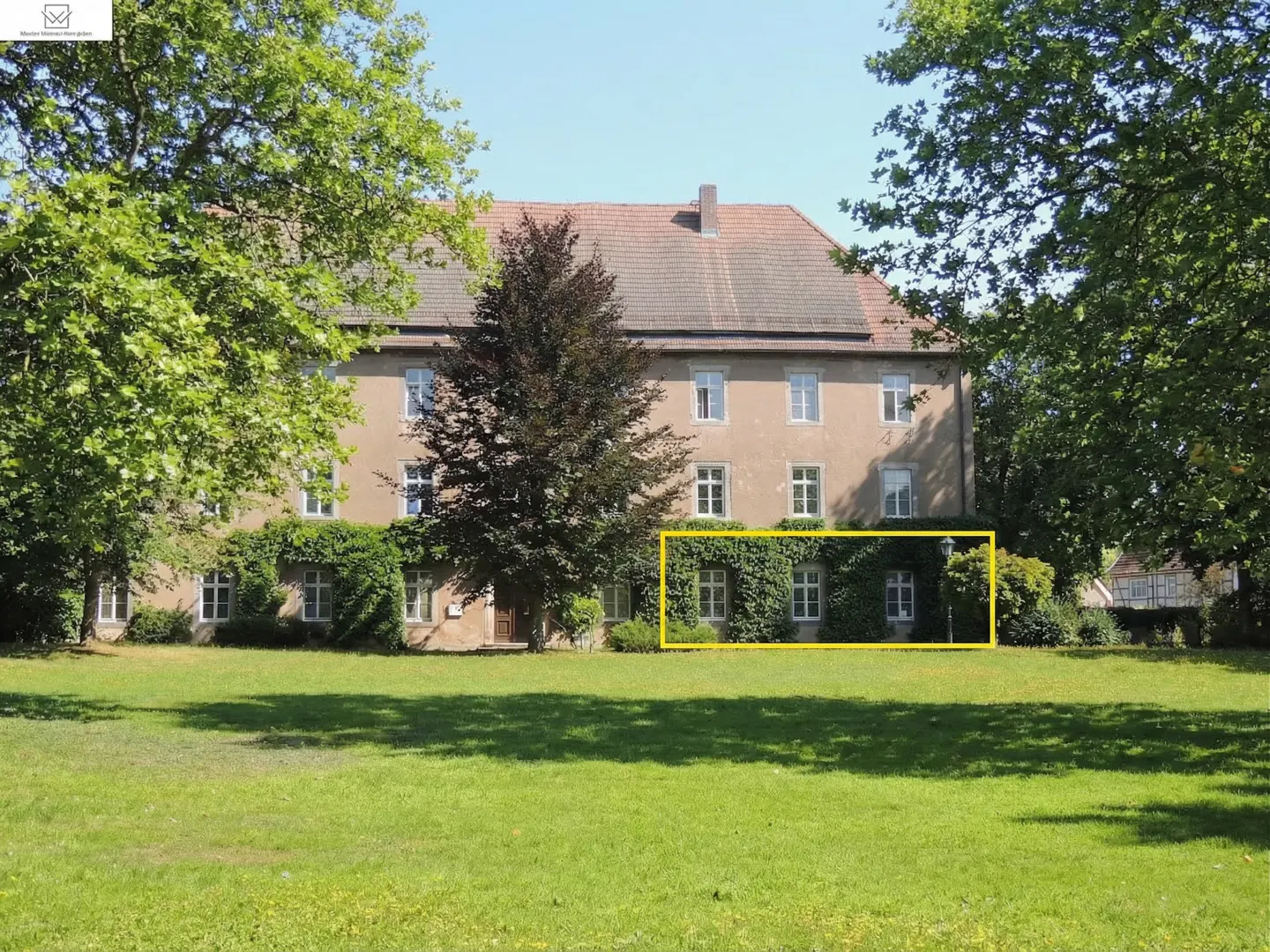 Exterior view of a beige two-story building with a red tile roof, surrounded by green trees and grass. Windows are framed by climbing ivy.