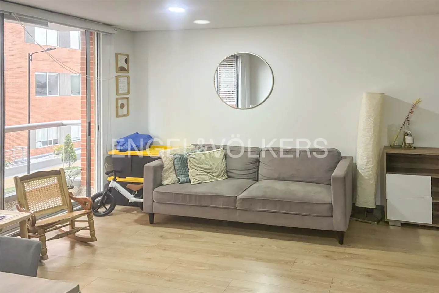 Living room with a gray sofa, wooden chair, round mirror, and a window with a view of a brick building.