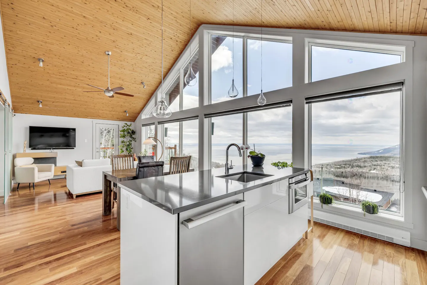 A modern kitchen with a white island, gray countertop, and large windows overlooking a scenic view. The room has wood floors and a wood ceiling.