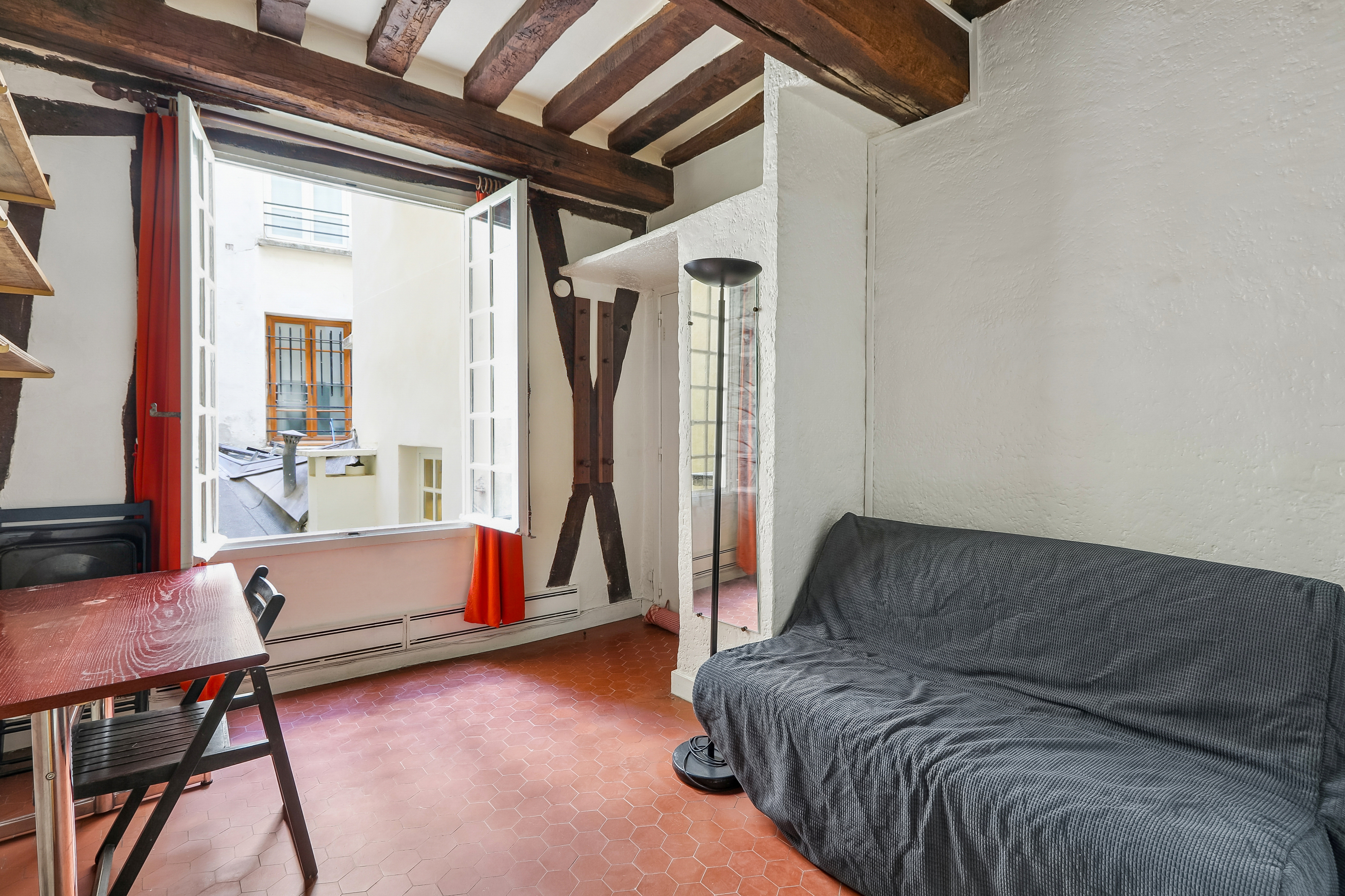 Living room with terracotta tile floor, exposed wooden ceiling beams, a grey sofa, and an open window overlooking a courtyard. [AI-generated text]