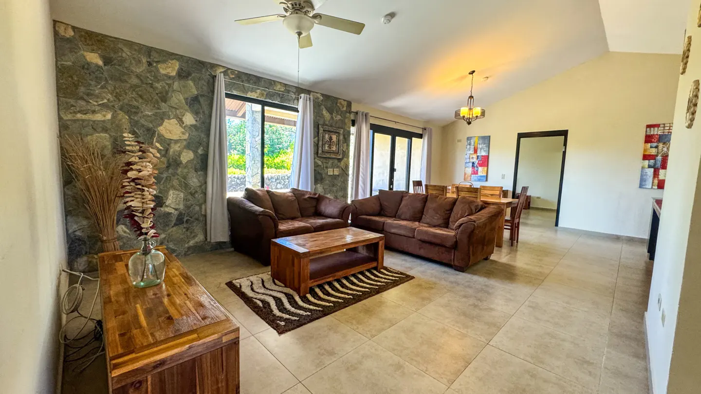 Living room with stone wall, brown sofas, wood table, and tile floor. A ceiling fan and light fixture are visible.