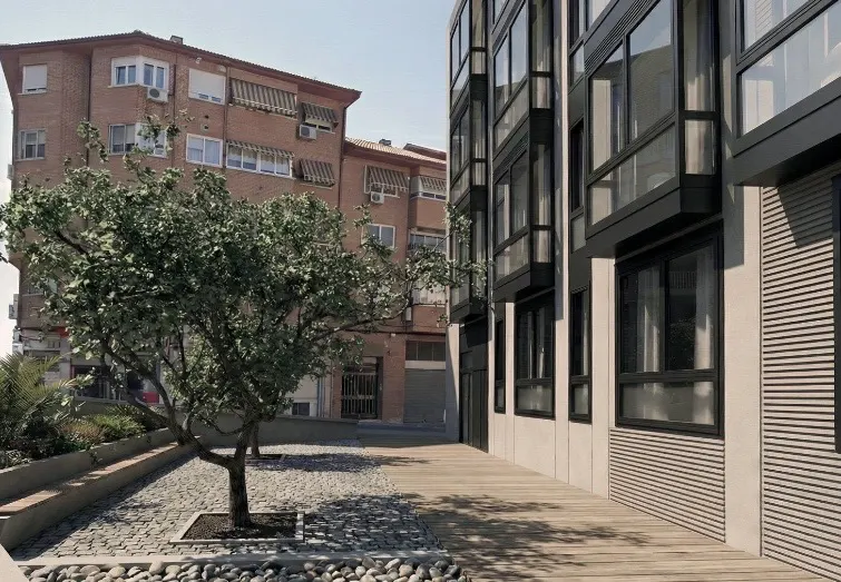 Exterior view of a modern apartment building with large windows, next to a brick building and a tree-lined courtyard.