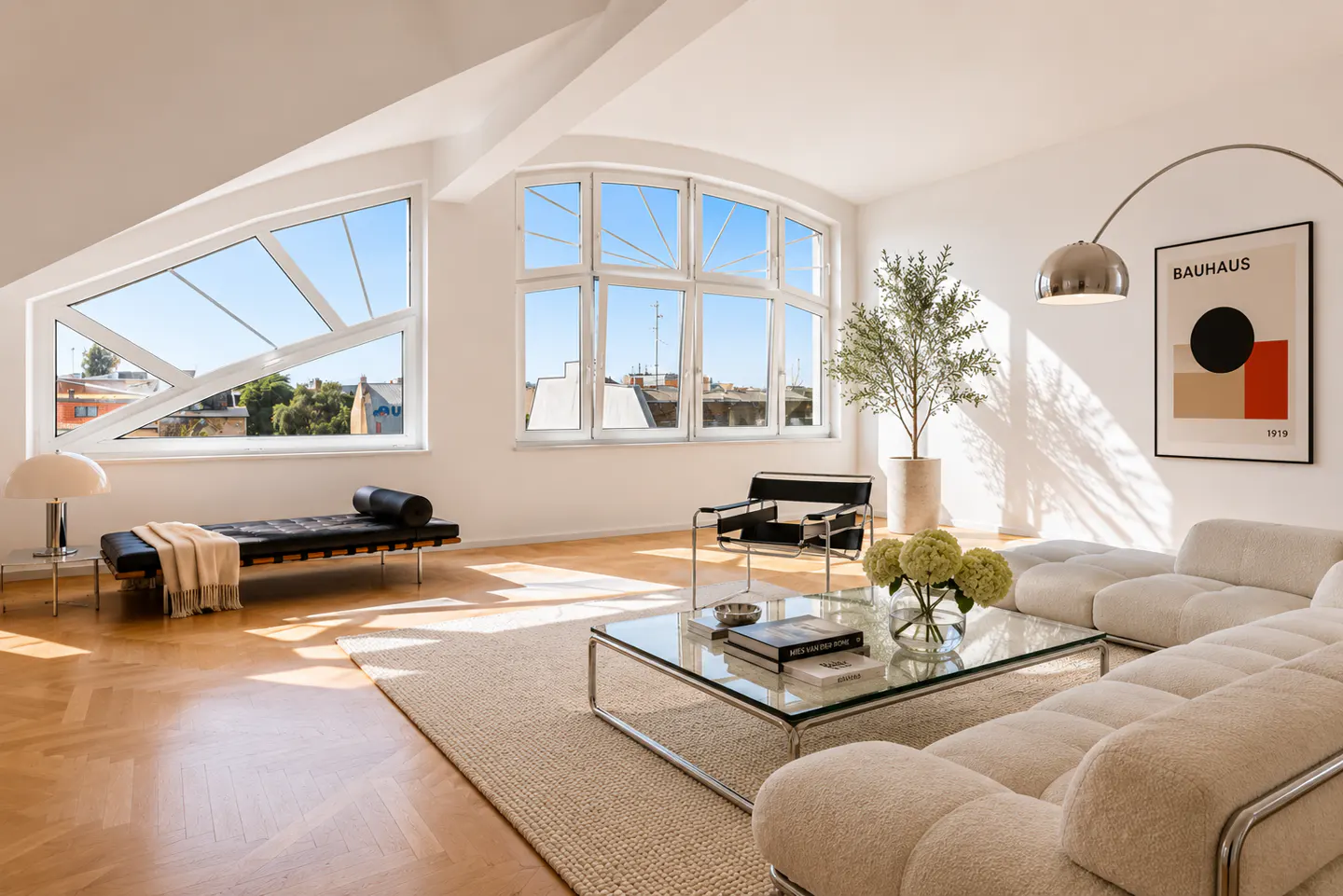 Bright, modern living room with large windows, a cream-colored sectional sofa, glass coffee table, and a Bauhaus art print on the white wall.