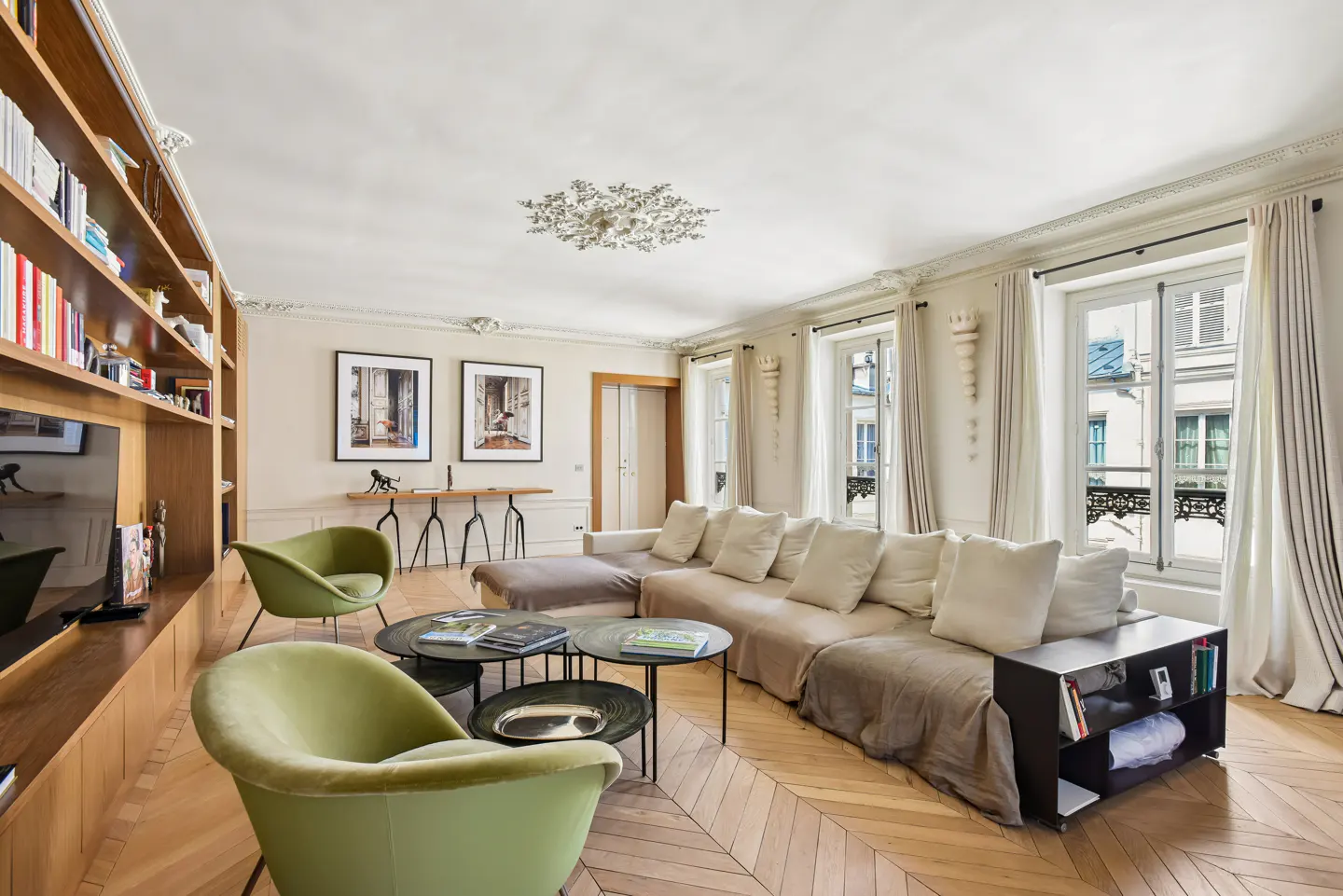 Bright living room with herringbone wood floors, a large beige sectional sofa, and two green armchairs. Built-in bookshelf and three windows with white curtains.