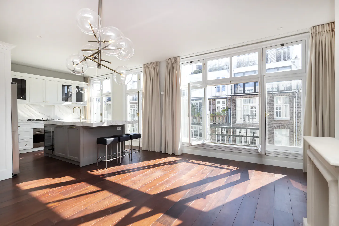 Bright, open-plan kitchen and living area with hardwood floors and large windows. A modern chandelier hangs above a gray kitchen island with black stools. Cream curtains frame the windows.
