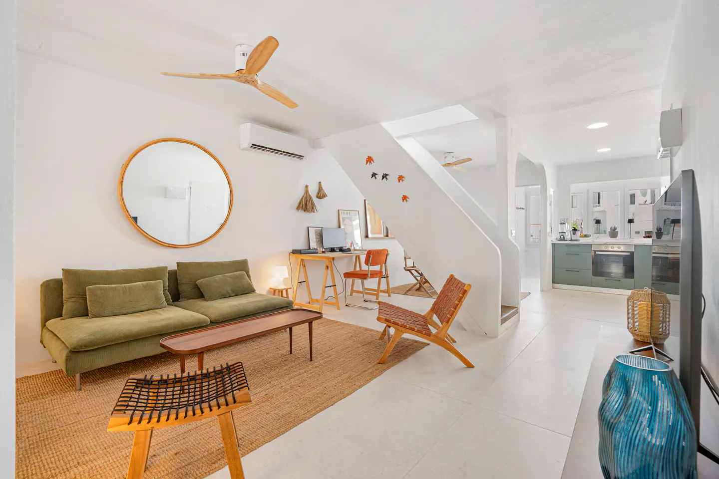 Bright, open-concept living space with white walls, a green sofa, and a jute rug. A wooden desk and chair sit near a staircase leading to a kitchen.