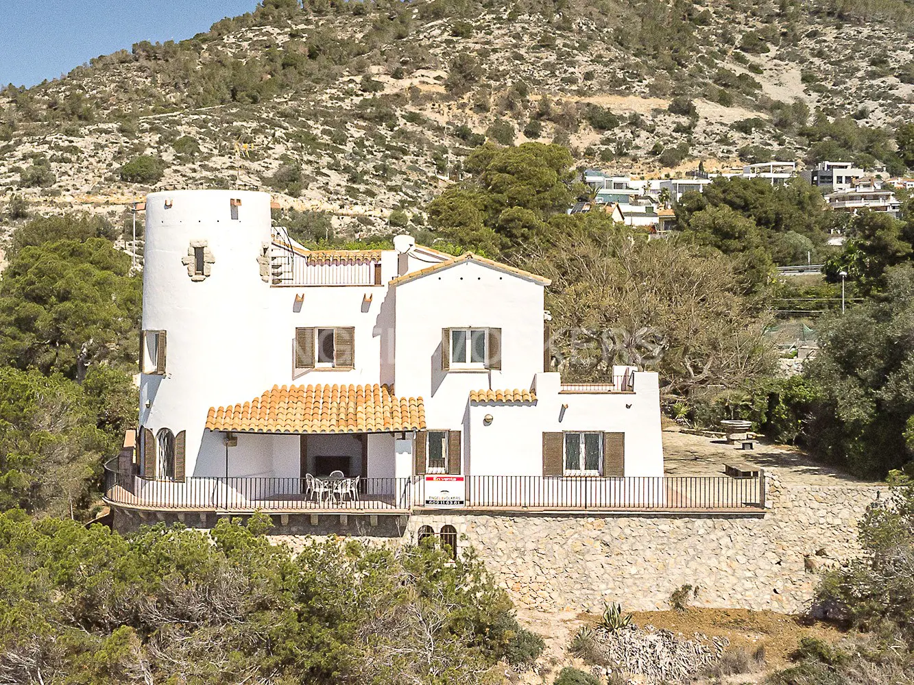 A white villa with a round tower and orange tile roof sits on a hill surrounded by green trees. A mountain is in the background.