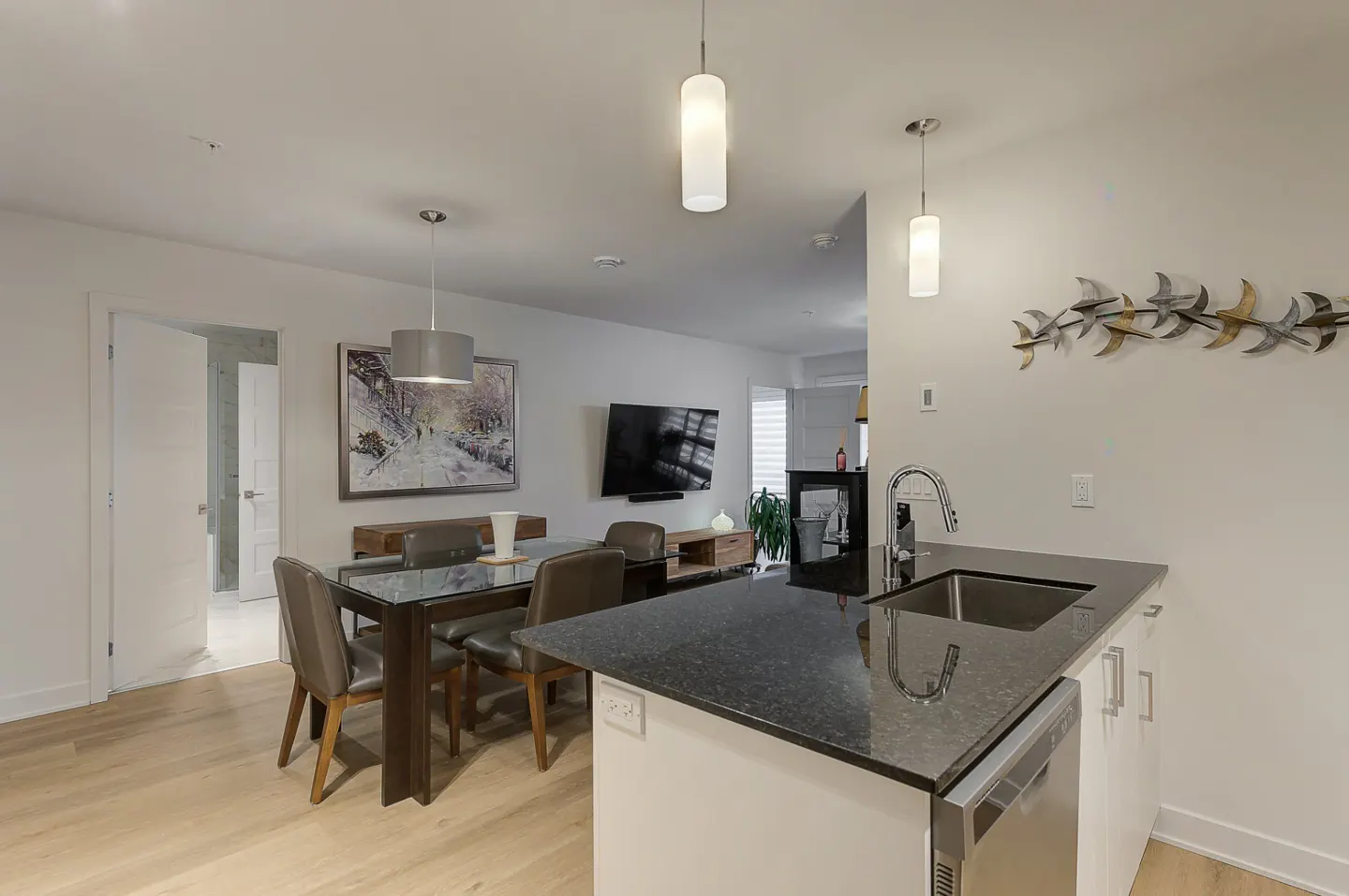 A modern open-concept living space with a kitchen island, dining table, and TV area. The kitchen has a black countertop and white cabinets.